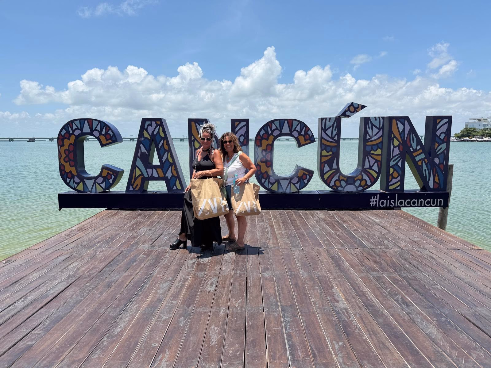 חופשה במקסיקו Two members of the group standing beside the colorful Cancun sign over the Caribbean water